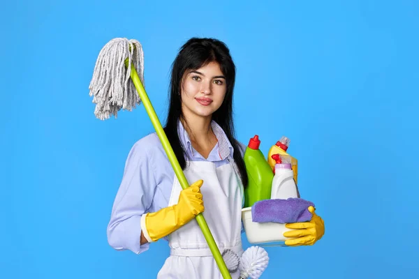 pretty woman in yellow rubber gloves and cleaner apron holding bucket of detergents and mop on blue background.
