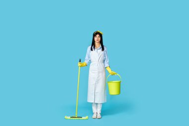 Lastik eldivenli bir kadın ve temiz önlüklü bir kadın mavi arka planda paspasla yerleri yıkıyor. Tam uzunluk