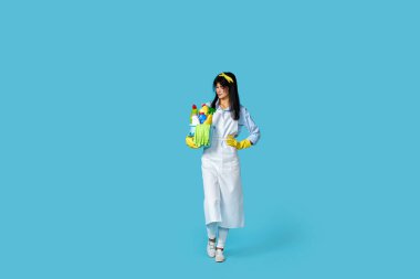 Profesyonel temizlikçi kadın lastik eldivenler ve temiz önlükle mavi arka planda bir kova deterjan tutuyor. Tam uzunluk