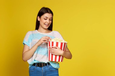 Mavi kazaklı güzel esmer kız sarı arka planda izole edilmiş patlamış mısır yiyor.