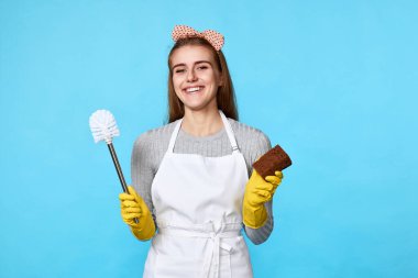 Lastik eldivenli mutlu kadın mavi arka planda sünger ve tuvalet fırçasıyla temizlik yapıyor..