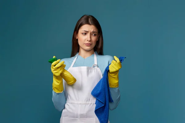 skeptic and nervous, frowning young woman in cleaner apron with cleaning rag and detergent sprayer on blue background.