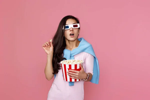 amazed woman watching horror movie holding bucket of popcorn isolated on pink background