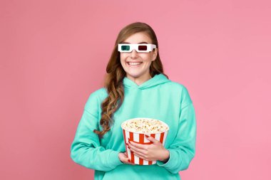 Sweatshirt 'lü gülen kıvırcık kadın pembe arka planda izole edilmiş patlamış mısır dolu bir kova tutuyor.