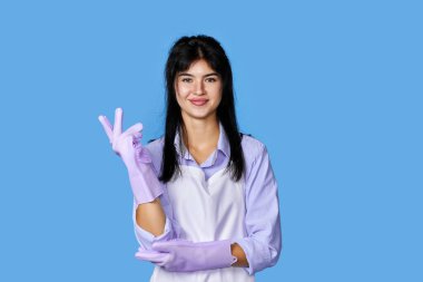 Lastik eldivenli ve temiz önlüklü güzel bir kadın mavi arka planda izole edilmiş kameraya bakıyor. temizlik