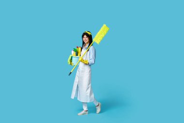 Lastik eldivenler ve temiz önlükle gülümseyen neşeli bir kadın. Temizlik malzemeleri ve mavi arka planda paspas.