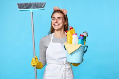 Sarı lastik eldivenli ve temiz önlüklü gülümseyen kadın elinde deterjan kovası ve mavi arka planda süpürge tutuyordu..