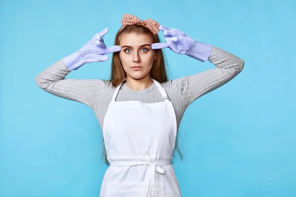 tired woman in gloves and cleaner apron standing with finger gun pointed to head on blue background.