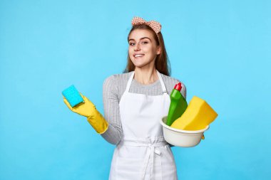 Lastik eldivenli, temiz önlüklü mavi süngerli ve mavi arka planda bir şişe deterjan olan genç bir kadın..