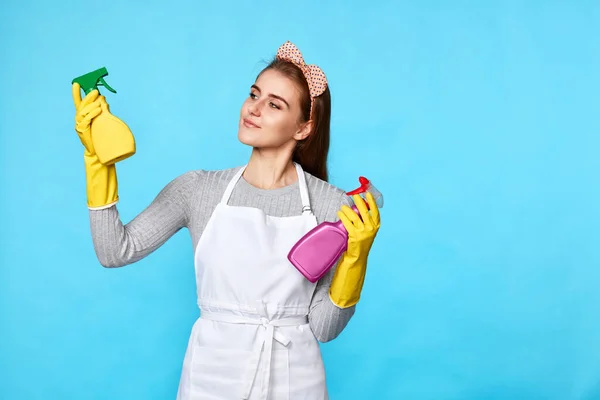 beautiful woman in yellow rubber gloves and cleaner apron chooses between two cleaning products on blue background.