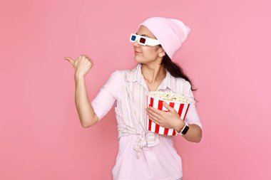 Şapkalı neşeli genç kadın, elinde patlamış mısırla pembe arka plandaki fotokopi alanını işaret ediyor..