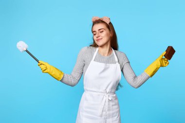Lastik eldivenli mutlu kadın mavi arka planda sünger ve tuvalet fırçasıyla temizlik yapıyor..