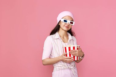 Cheerful young woman in hat watching movie film, holding bucket of popcorn on pink background.