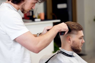 Berber dükkanındaki sakallı yakışıklının saçını kırpıcı ile kesiyor..