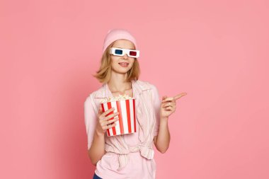 smiling woman in t-shirt and hat with popcorn pointing at the copy space on pink background.