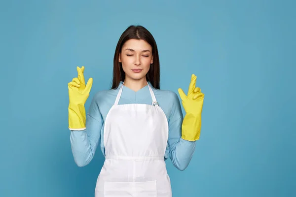 beautiful brunette caucasian girl in yellow rubber gloves with finger crossed gesture isolated on blue background.