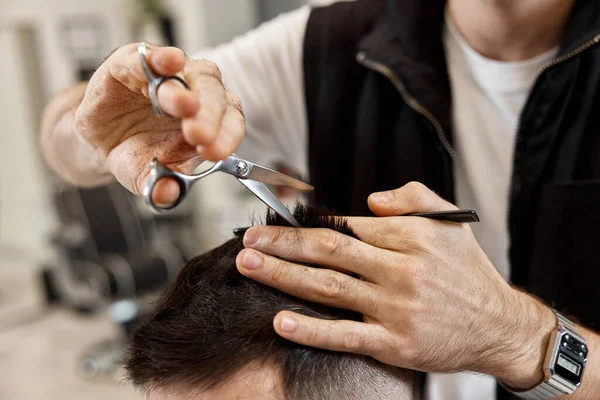 Professional hairdresser does haircut for caucasian bearded man using comb and scissors at barber shop.