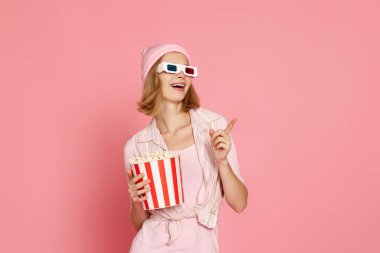 smiling woman in t-shirt and hat with popcorn pointing at the copy space on pink background.