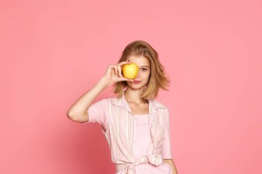 blonde woman covers one eye with fresh apple on pink background.