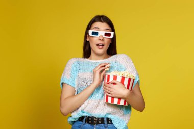 terrified brunette woman in 3d glasses watching horror movie , holding bucket of popcorn on yellow background