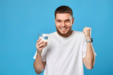smiling handsome bearded man in white t-shirt with raised arm holding glass of water on blue background