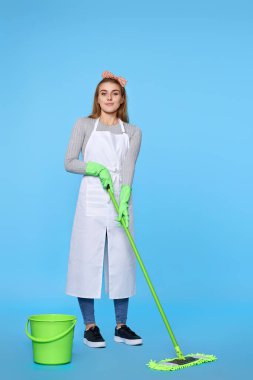 young attractive woman in green rubber gloves washing floor with mop on blue background. Full length