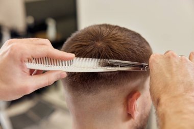 Professional hairdresser does haircut for caucasian bearded man using comb and scissors at barber shop. close-up