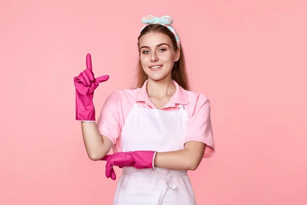 beautiful woman in rubber gloves and apron holding index finger up with new idea on pink background. cleaning