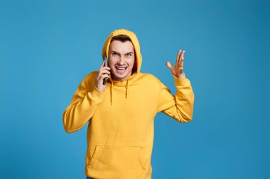 Sarı kazaklı endişeli bir adam cep telefonu tutuyor ve mavi arka planda öfkeli bir şekilde izole edilmiş hissediyor.