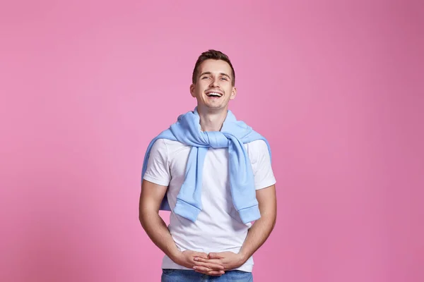 smiling young guy in white t-shirt and blue sweatshirt posing on pink studio background