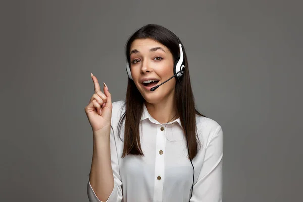 beautiful woman with headset holding index finger up with new idea and looking at camera on gray studio background. Call center, business people concept