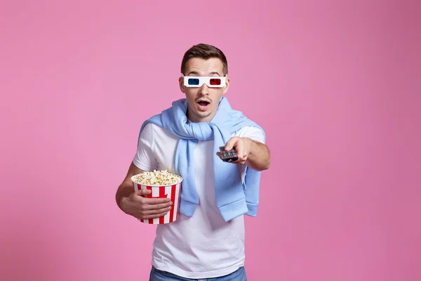 amazed man in 3d glasses using remote controller watching movie with big bucket of popcorns on pink background