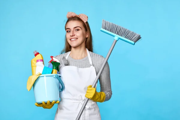 young happy woman in yellow rubber gloves and cleaner apron holding bucket of detergents and broom on blue background.