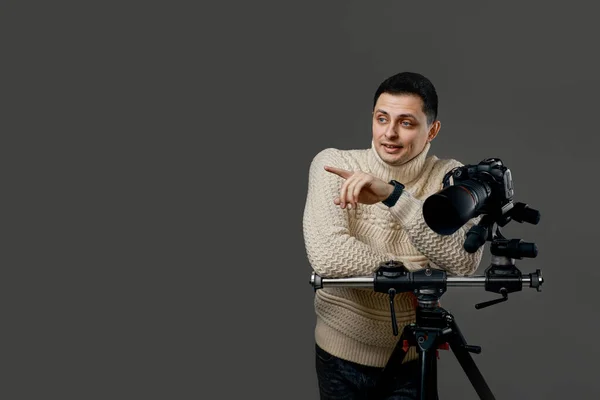 Professional young handsome photographer in sweater with digital camera on tripod pointing finger at the side on gray background. copy space