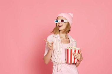 smiling woman in t-shirt and hat with popcorn pointing at the copy space on pink background.