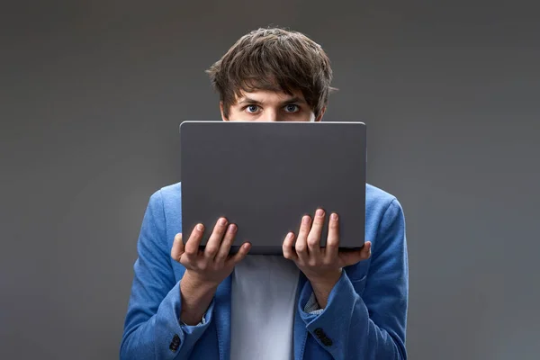 caucasian young guy hiding his face with laptop on gray background