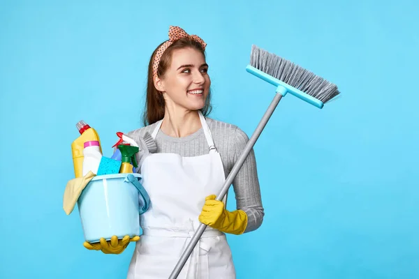 smiling woman in yellow rubber gloves and cleaner apron holding bucket of detergents and broom on blue background.