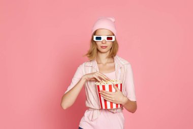 3 boyutlu gözlüklü çekici sarışın kadın pembe arka planda izole edilmiş patlamış mısır kovası tutuyor.