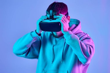 man in sweatshirt using virtual reality headset on blue background. Neon lighting