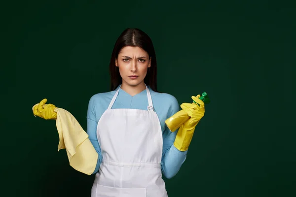 skeptic and nervous, frowning woman in gloves and cleaner apron with cleaning rag and detergent sprayer on green background.