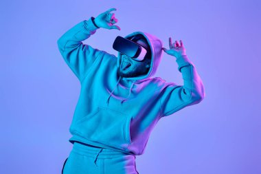 man in sweatshirt using virtual reality glasses and playing video games on blue background. Neon lighting