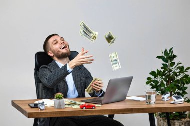 young bearded businessman working on laptop and throwing money while sitting on chair at desk. banknotes fly in the air