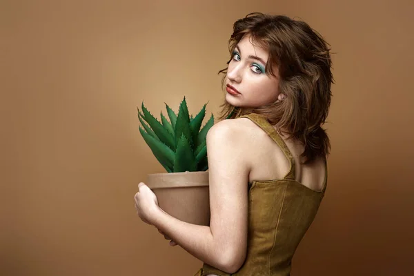 young woman with stylish hairstyle posing with plant in pot on beige background. copy space