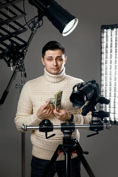 Professional handsome photographer with digital camera on tripod holding hundred dollar bills on gray background with lighting equipment