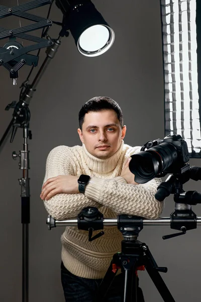 Professional photographer with digital camera on tripod looking at the camera on gray background with lighting equipment