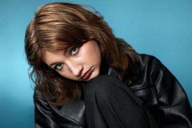 young woman with stylish hairstyle posing in black leather jacket on blue background.
