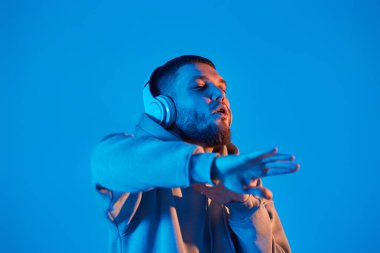 handsome bearded man with headphones in sweatshirt enjoying favorite music on blue neon background. Neon lighting
