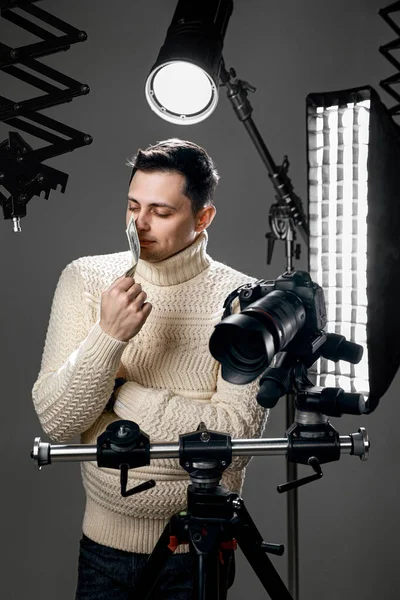 专业英俊的摄影师 三脚架上有数码相机 带着照明设备 在灰色背景下闻到钱的味道 — 图库照片