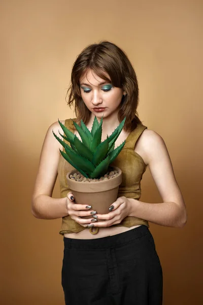 young woman with stylish hairstyle posing with plant in pot on beige background.
