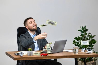young bearded businessman working on laptop and throwing money while sitting on chair at desk. banknotes fly in the air